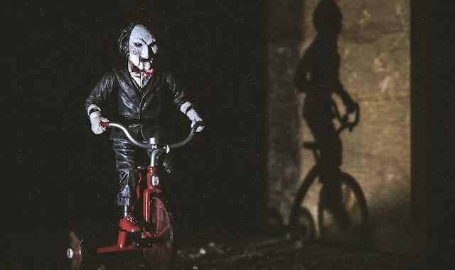 A ventriloquist puppet on a tricycle on the left side of the photo. The puppet has short black hair, white face with red lips and red spiral pattern blush on its cheeks. The doll is also wearing a black suit with a white button up underneath and a red bowtie. Light from the left is shining on the puppet which casts a stark shadow on a concrete wall to the right of the puppet. Everything behind the doll is pitch black.